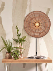 Good&Mojo Wandleuchte Tanami aus Rattan und Eisen in Natur mit rundem Rattanschirm, eingeschaltet über einem Holztisch.