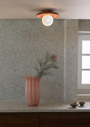 Wand- und Deckenleuchte Tivoli von RoMi in Terrakotta mit weißem Glasschirm, darüber eine rosa Vase mit Kiefernzweig vor Terrazzowand.