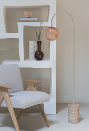 It’s about RoMi Stehleuchte Tivoli in Sand mit Glasschirm und Travertinfuß, neben einem Sessel mit Holzgestell und Wandregal.