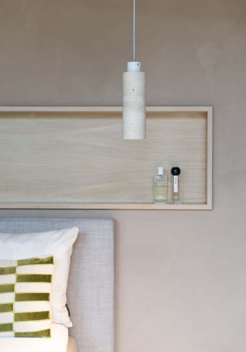 Pendelleuchte Tivoli aus weißem Travertin mit Zylinderschirm, über einem Holzregal an einer Wand hängend.