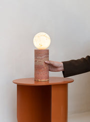 Tischleuchte RoMi Tivoli in Terrakotta mit zylindrischem Travertin-Fuß und leuchtendem Glasschirm auf einem Beistelltisch.