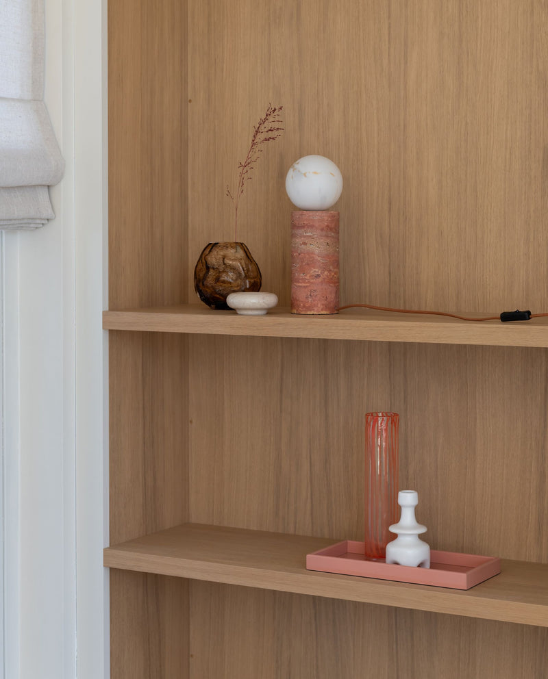 Die Tischleuchte "It's about RoMi Tivoli" aus terrakottafarbenem Travertin mit weißem Glasschirm steht auf einem Holzregal.