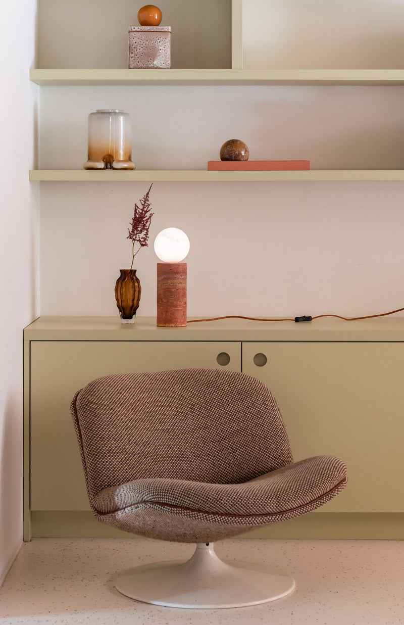 Die Tischleuchte Tivoli von RoMi aus Marmor und Glas in Terrakotta mit Travertin-Fuß und Glaskugelschirm auf einem Sideboard.