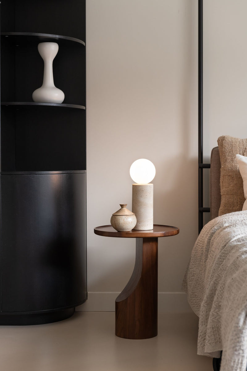 Tischleuchte Tivoli aus Travertin mit weißer Glaskugel als Schirm, auf einem Beistelltisch aus dunklem Holz neben einem Bett.