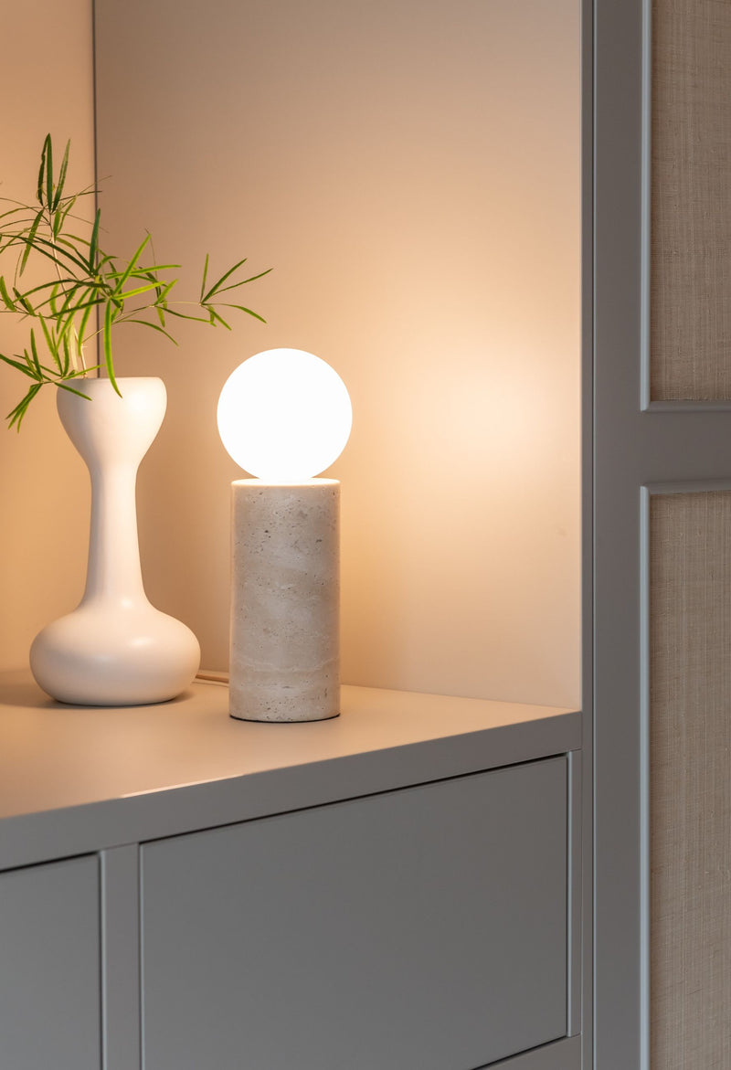 Tischleuchte Tivoli von It's about RoMi aus Travertin mit leuchtendem Opalglas-Schirm auf einem grauen Sideboard.