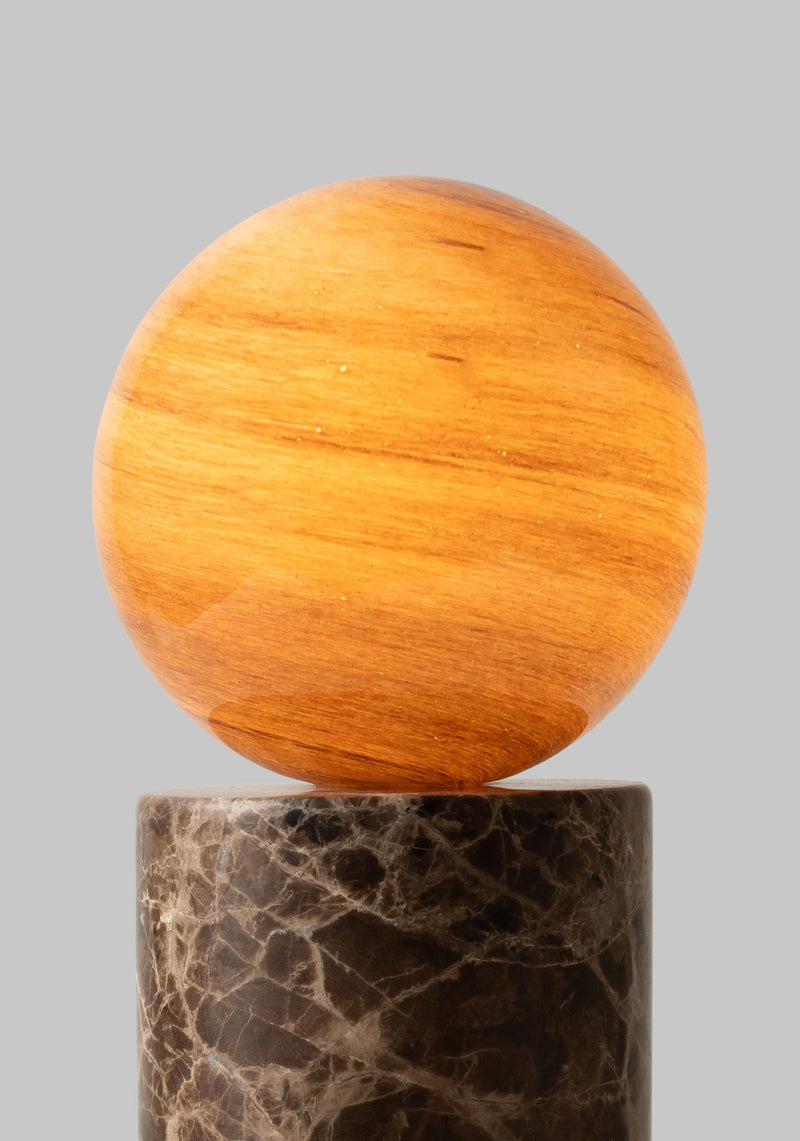 Eine goldbraune Glaskugel mit feiner Maserung auf einem zylindrischen Sockel aus braunem Marmor vor grauem Hintergrund.