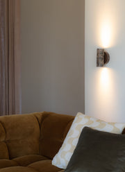 Die Wandleuchte Tivoli von RoMi aus Marmor in Braun leuchtet an einer hellen Wand über einem braunen Samtsofa.