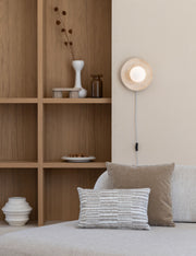 Wandleuchte Tivoli mit rundem Travertin-Schirm und opalweißer Glaskugel, neben Holzregal und Sofa.