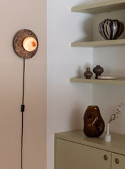 Die RoMi Wandleuchte Tivoli aus braunem Marmor und Glas leuchtet mit eingeschaltetem Glasschirm an der Wand, mit schwarzem Kabel.