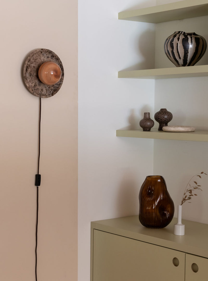Die Wandleuchte RoMi Tivoli in Braun: runde Marmorscheibe mit gläsernem Schirm an der Wand, daneben Keramikvasen.