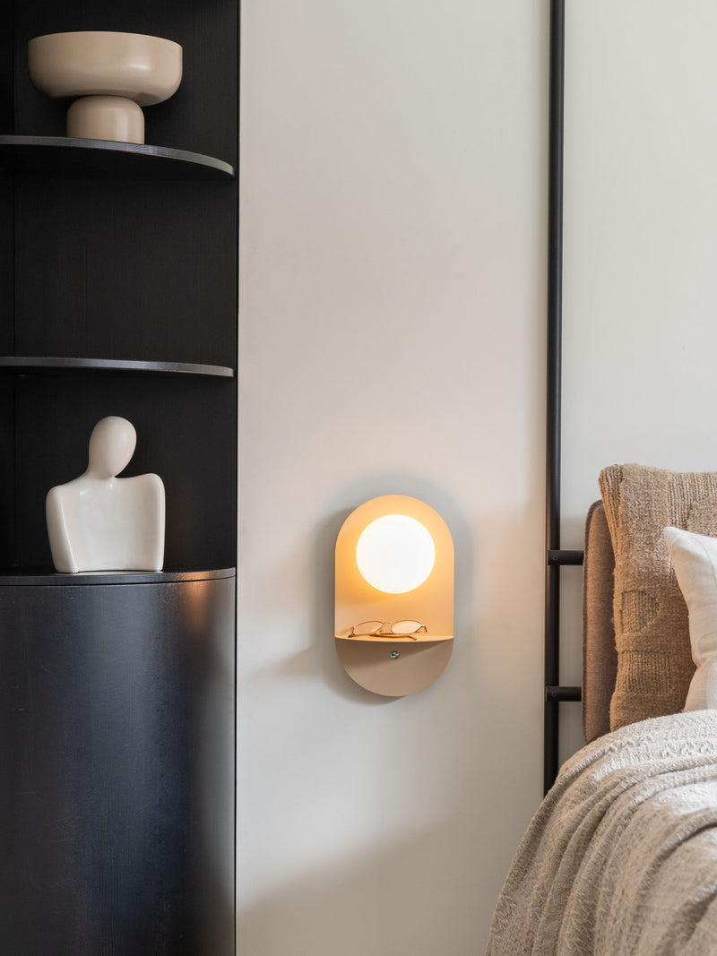 Die Wandleuchte RoMi Tokyo: beige Wandleuchte mit rundem Glasschirm und Ablage, eingeschaltet im Schlafzimmer montiert.