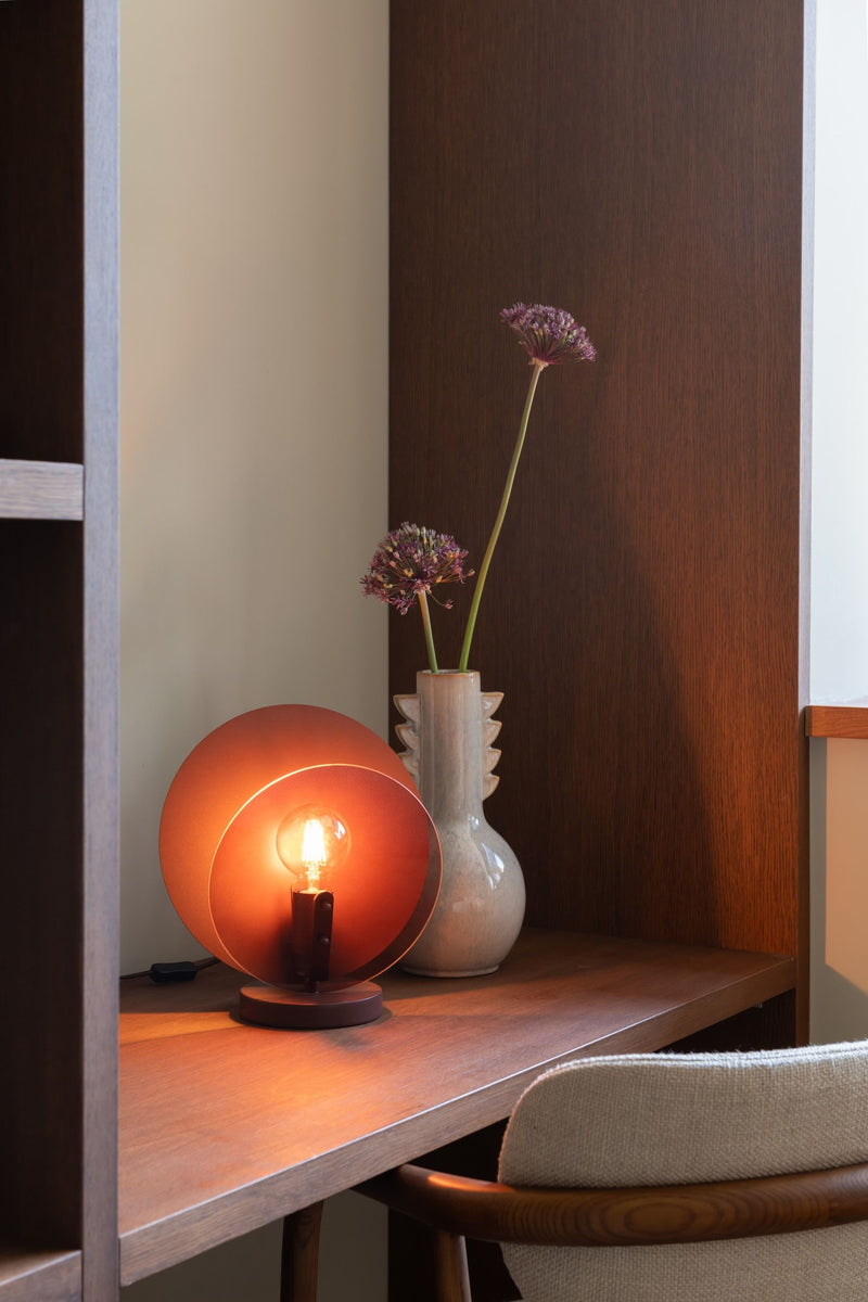 Tischleuchte „It's about RoMi Toledo" mit rundem, braunem Glasschirm und sichtbar brennender Glühlampe auf einem Holztisch.