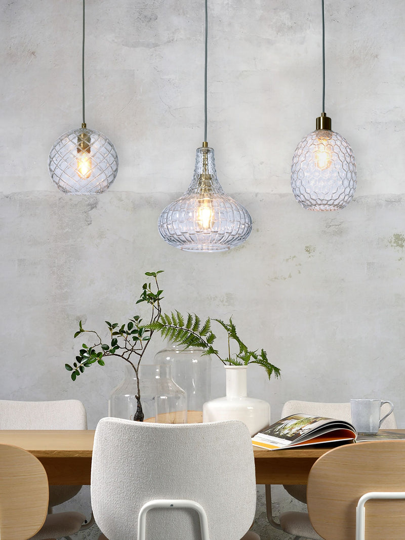 Drei klare Glas-Hängeleuchten mit gemustertem Schliff und Messingfassung über einem Holztisch mit Stühlen und Vasen.