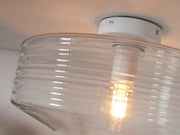 Detailansicht der Deckenleuchte It's about RoMi Verona mit klarem, geripptem Glasschirm und weißem Metallgestell, daneben eine leuchtende Glühlampe.