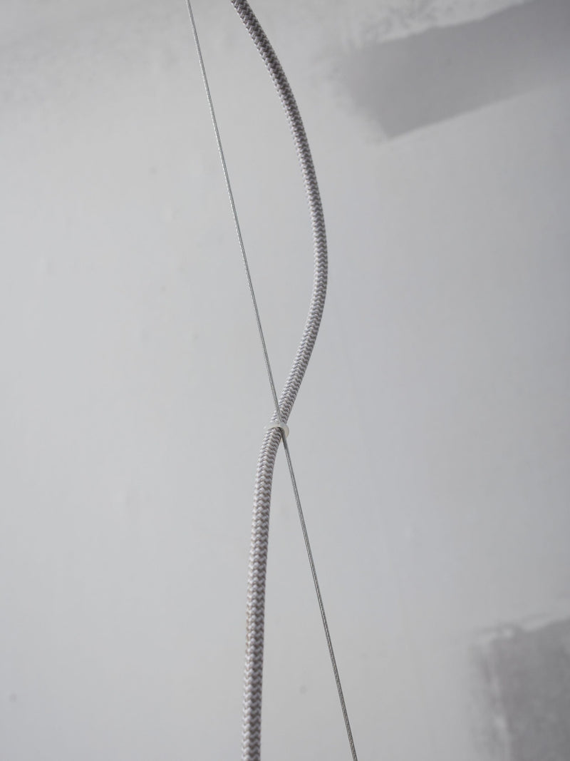 Detailaufnahme eines grau-weiß geflochtenen Hängeleuchten-Kabels, das an einem Verbindungsstück über einer hellen Wand zusammenläuft.