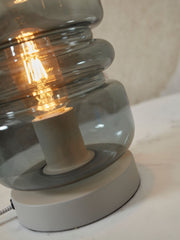 Detailansicht der Tischleuchte Verona von It's about RoMi: gestapelter grauer Glasschirm mit leuchtender Glühlampe auf hellgrauem Sockel.