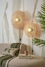 Zwei runde Wandleuchten aus Rattan mit Eisengestell an heller, strukturierter Wand, die warmes Licht abstrahlen.