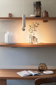 Die Hängeleuchte RoMi Milan in Sand hängt über einem Holzregal mit weißer Vase und Glasvasen, die warm strahlt.