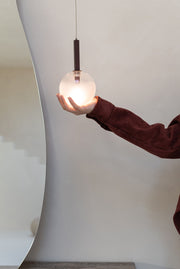 Eine Hand mit bordeauxrotem Ärmel hält die Hängeleuchte Murano mit mattiertem Glasschirm, deren Lampe leuchtet.