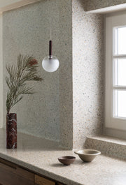 Hängeleuchte aus mattiertem Glas mit bordeauxfarbenem Eisengestell über einer Terrazzo-Arbeitsplatte, daneben eine Vase mit Zweig.