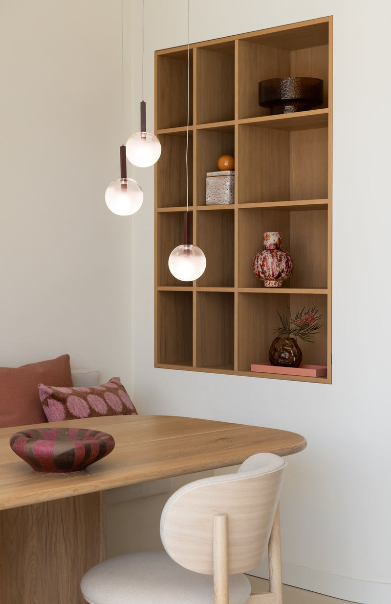 Drei weiße Glaskugel-Hängeleuchten mit bordeauxroten Eisenstangen über einem Holztisch mit Sitzbank.