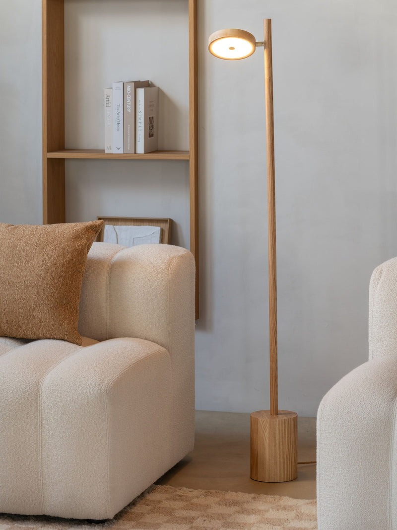 Stehleuchte Okinawa aus Eschenholz mit rundem LED-Schirm, daneben ein cremefarbenes Sofa und ein Holzregal.