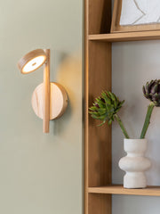 Good&Mojo Wandleuchte Okinawa aus Eschenholz: runder Holzschirm mit LED-Licht vor einer sagegrünen Wand.