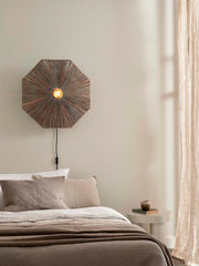 Good&Mojo Wandleuchte Panama aus Raffia: achteckiger, braun gewebter Schirm mit Lichtquelle in der Mitte, über einem Bett.
